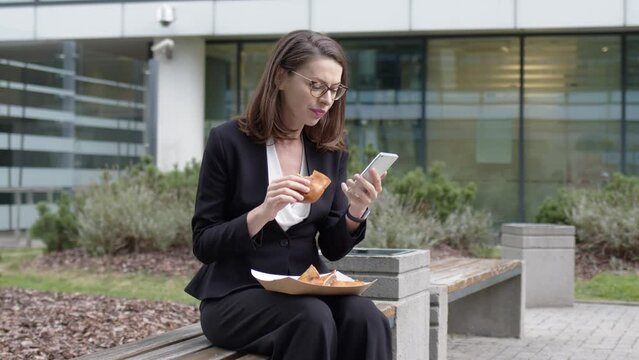 Businesswoman browsing smartphone and snacking with sandwich in yard