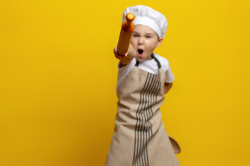 a funny boy cook in an apron and hat holds a rolling pin in his hands and shouts to attack on an...