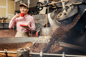 A happy female factory worker smelling fresh cup of coffee while standing next to a roasting machine.