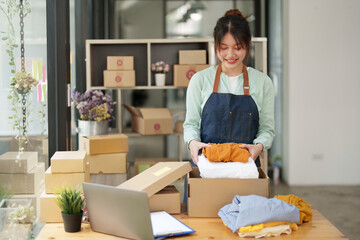 Portrait of a small startup Asian female entrepreneur SME owner picking up a yellow shirt before packing it in an inner box with a customer. Online Business Ideas and Freelance