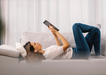 A woman with earphones lies in the living room and reads an online book.