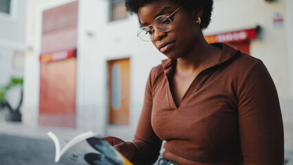 African woman wearing glasses sitting on the street with book. F
