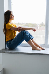 Young woman looking through the window with a city view, sitting on a windowsill
