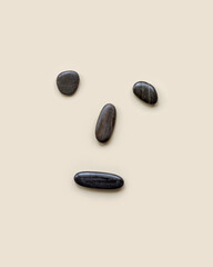 Anthropomorphic face from black colored sea stones on beige background, frowning, unhappy, sad mood, sad expression. Summer creative flat lay, minimal style, top view