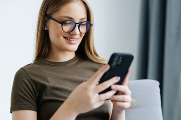 Beautiful young woman holding phone and looking at camera with smile while sitting in big comfortable chair at home