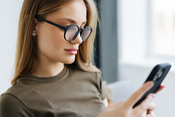 Beautiful young woman holding phone and looking at camera with smile while sitting in big comfortable chair at home