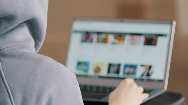 Rear view unknown hacker young programmer web designer in hood look at laptop display develop website scroll page choose trailer film use internet for job search information online construct software