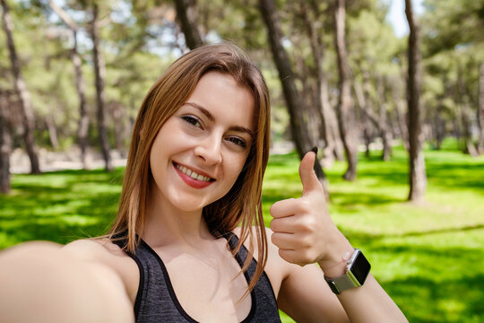 Happy brunette woman wearing black sports bra standing on city park, outdoors showing thumb up and taking selfie photo. Looking at camera.