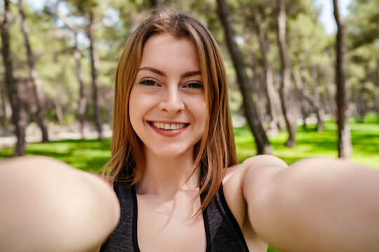 Cute brunette woman wearing sports bra standing on city park, outdoors taking selfie on front camera with two hands enjoying break having fun video call with friend.