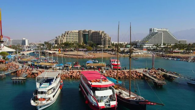 Aerial Shot Of People Walking On Footpath At Harbor, Drone Flying Backwards Over Red Sea During Sunny Day - Eilat, Israel