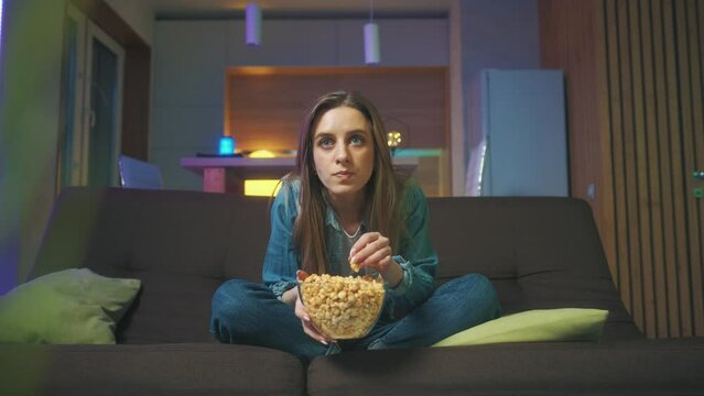 A shocked young girl with popcorn is watching a tv program or movie at night. Unexpected turn of events in a film. The amazed woman is popeyes surprised watching tv sitting on a couch at home.