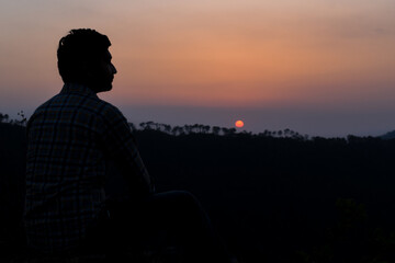 Fototapeta premium Guy sitting looking sideways silhouette pose and sun setting behind mountain chain in background