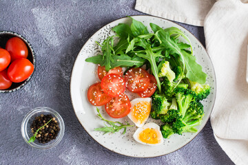 A plate with broccoli, arugula, tomato and boiled egg on a plate on the table. Top view