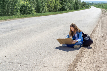A young girl is sitting on the side of the road and writing on cardboard while hitchhiking around the country. The girl is trying to catch a passing car for a trip. A girl with a backpack went on