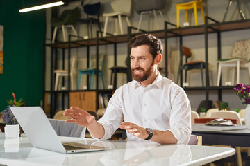 Front view of happy designer, architect sitting at table, working, using laptop. Brunette male with beard looking at screen, smiling, designing. Concept of urban lifestyle.
