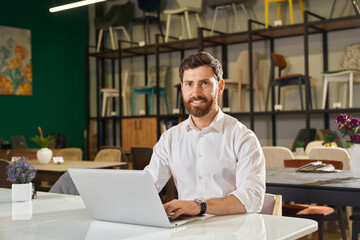 Front view of handsome designer sitting at table, using laptop. Brunette man with beard wearing...