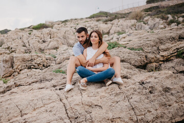 Naklejka na ściany i meble A Hispanic man is holding between his legs his brunette Latina girlfriend while both are sitting on the rocks in Spain. A man is hugging his wife on a date on the coast at sunset in Valencia.