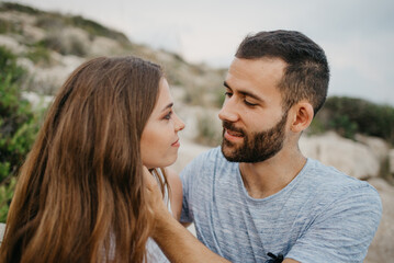 A close photo of a Hispanic man who is stroking with his hands the face of his Latina girlfriend on the rocky sea coast in Spain. A couple of tourists in the evening on the hills in Valencia