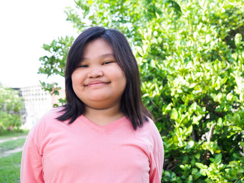 Portrait Of A Chubby Asian Girl. Cute, Beautiful, Black Hair, 10 Year Old, Wearing A Pink Shirt. Stand Smiling Happily And Freely. With Bright Eyes In Front Of The Wall With A Park Background Thailand