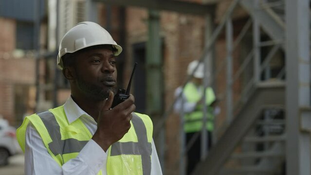 Responsible contractor in hardhat using walkie talkie while standing near outdoor building site. African american man managing working process to avoid construction damages.