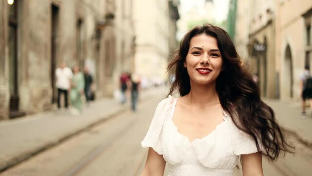 Smiling woman in white dress walking down the city street. Flirty girl with long dark hair turning around in city centre.