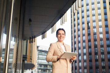 Portrait of cheerful satisfied middle-aged brunette businesswoman in jacket standing in business district and using tablet for remote work