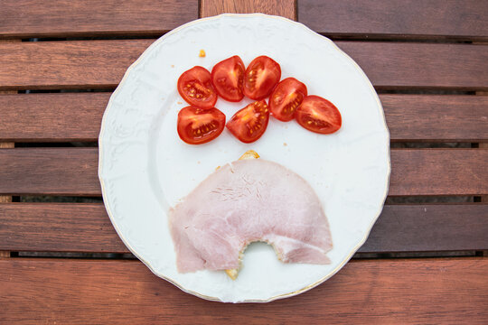 Snack on a white plate on a wooden table. Red tomatoes and bitten toat with ham and butter.