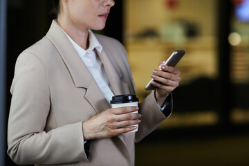 Close-up of pensive woman in jacket drinking coffee from takeout cup and reading sms on phone