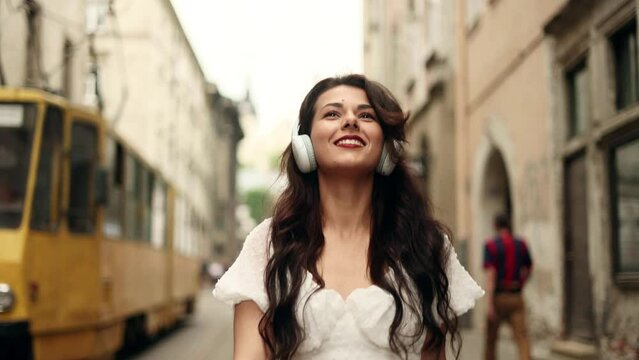 Closeup face of beautiful young girl with long hair. Slow motion of smiling young girl listening to music in headphones at the city street. 