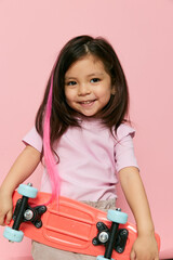 a cute little girl is standing on a pink background in a pink T-shirt, holding her skate in one hand in front of her, and raising the other behind her head