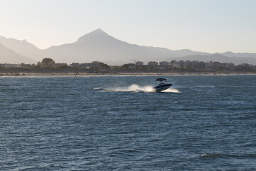 Blue and white speedboat creates white waves on the sea against the backdrop of a beach filled with tourist and moutains in  the distance.