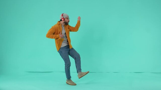 Happy emotional male wearing eyeglasses and headphones moving to rock music and imagining playing bass guitar against blue studio background. Cheerful rock fan listening to music during dance