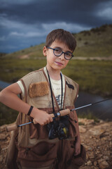 An elementary boy with glasses wearing a fishing vest and holding a fishing rod.
