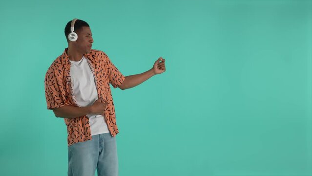 Positive african american guy listening to rock music in headphones and pretending to play bass guitar during dance. Cheerful dark-skinned man enjoying dance and music isolated against blue background