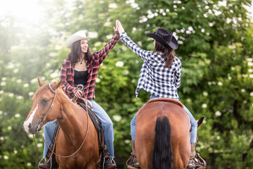 Two cowgirl friends with horses facing the opposite direction give a high five