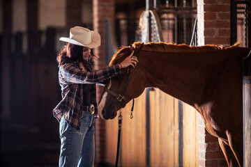 Young cowgirl takes bridle off the horse's head inside the stable after a ride.