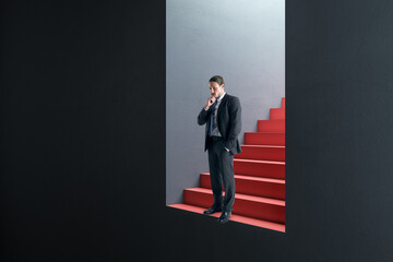 Attractive thoughtful young european businessman standing in abstract concrete wall opening with red stairs and mock up place. Door, opportunity and success concept.