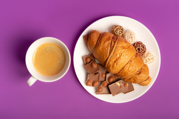 Sweet breakfast with morning hot coffee, tasty french croissant and sweet milk chocolate with nuts on white round plate from above on violet background. 