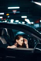 vertical photo from the side, at night, of a woman sitting in a black car and looking out of the window looking into the side view mirror holding her hand near her face