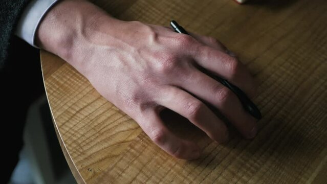 Fit male hand with pen knocking fingers on the table nervous and waiting for someone. Close up of anxious hand tapping wooden surface. Concept of delaying business meeting due to partner being late
