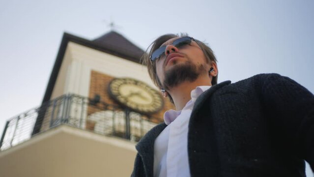 Confident successful crypto trader in sunglasses looking at the watch waiting for the meeting. Handsome young businessman standing under the clock tower and checking his watch. Successful rich male