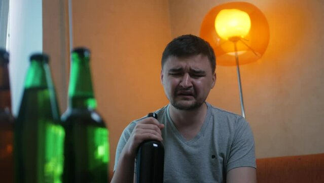 Upset young guy sitting on the sofa and drinking alcohol after he broke up with a girl. Crying depressed man with alcohol abuse consumes wine at home. Male in a terrible mood drinking after bad day
