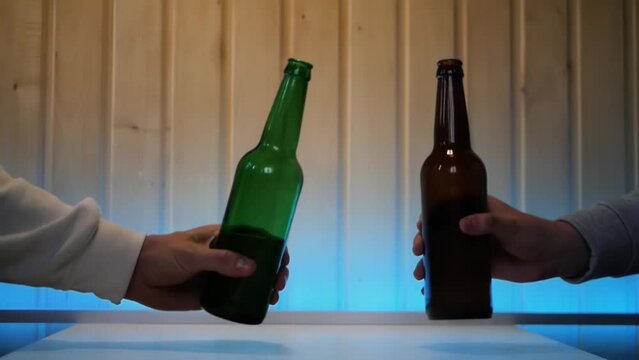 Close up of two male hands clinking a pair of beer bottles in greeting. Two men cheer beers at home in a pleasant atmosphere with blue blacklight. Concept of a toast to friendship with beer drinks