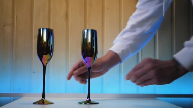 Close up of white male hands serving the table with fancy glasses and a bottle of wine. Waiter putting two glasses and a bottle of alcohol preparing for romantic dinner. Preparation of table for date
