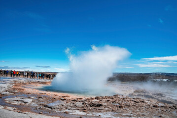 Tourists looking at eruption of Strokkur geyser. Smoke emitting from landscape against blue sky. Men and women exploring famous attraction in valley during sunny day.
