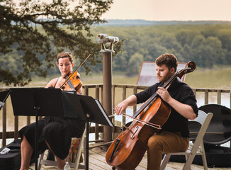 Female violin player and male cello player playing duet on wooden deck over Missouri River on...