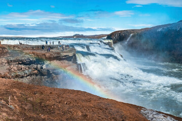 Tourists enjoying view of Gullfoss waterfall in Golden Circle. Scenic view of falling water with rainbow against blue sky. People exploring natural scenery in valley.