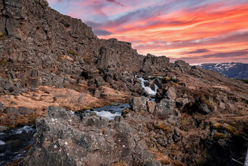 Idyllic view of Drekkingarhylur by rocky cliffs during sunset. Beautiful waterfall amidst rock formations against cloudy sky. Scenic tourist landmark at Thingvellir National Park.