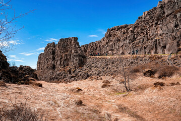 Fototapeta premium Tourists exploring beautiful volcanic landscape. People are standing by rock formation against sky during sunny day. Concept of tourism at dramatic land in northern Alpine region.