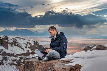 Male tourist using laptop while sitting on snow covered rocky cliff. Scenic view of volcanic landscape during winter season. Young explorer using wireless technology against cloudy sky.